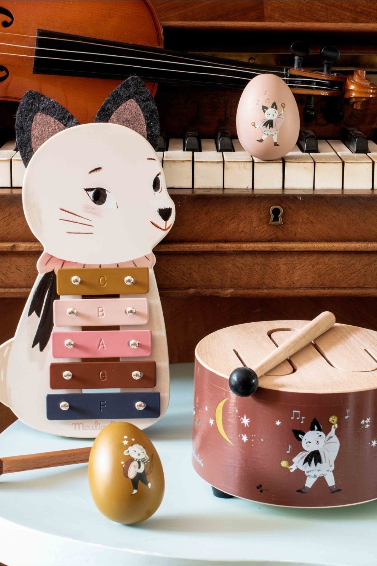 A children's wooden toy set featuring a cat-shaped xylophone, a small wooden drum with a cat illustration, matching maracas, and a toy mallet, styled in front of a piano with a violin in the background.

Moulin Roty -  Après la Pluie - Xylophone - Strawberries & Cream - Ingrid Lewis - Designer